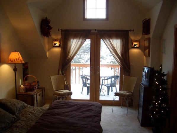A bedroom with a large bed, a desk with a lamp, a chair, and a sliding glass door leading to an outdoor patio with chairs and a table.