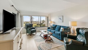 A spacious living room with a large flat-screen TV, a blue sofa, a leather armchair, and a coffee table. - Beautiful First Floor Oceanfront Condo at Amelia Surf and Racquet-Close to Pool! (Fernandina Beach)