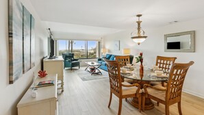 A spacious living area with a dining table set for four, a sofa, and a large window offering a view of the city. - Beautiful First Floor Oceanfront Condo at Amelia Surf and Racquet-Close to Pool! (Fernandina Beach)