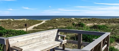 Un banco de madera en una terraza de madera con vistas a una playa, dunas y cielo despejado.
