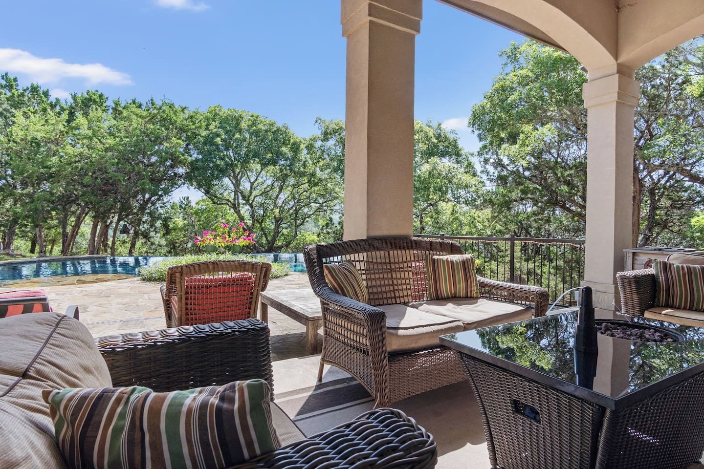 An outdoor patio with wicker furniture and a glass table, surrounded by trees and a clear sky.