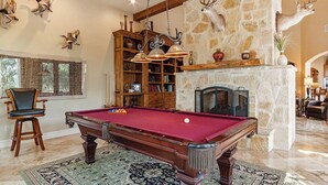 A pool table in a room with a stone fireplace, wooden shelves, and mounted animal heads. - Rockin B Summit | Luxury Retreat with Pool & Creek (Pipe Creek)