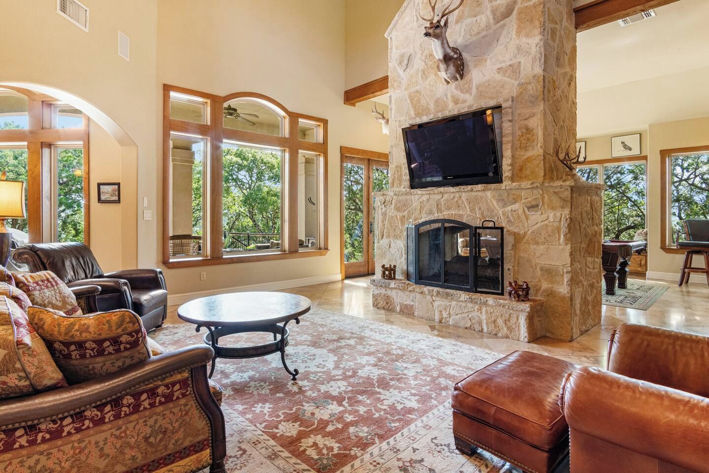 A spacious living room with a stone fireplace, leather furniture, and large windows offering a view of the outdoors.