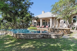 A pool with a stone border, a covered patio with chairs, and a house with columns. - Rockin B Summit | Luxury Retreat with Pool & Creek (Pipe Creek)