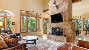 A spacious living room with a stone fireplace, leather furniture, and large windows offering a view of the outdoors. - Rockin' B Ranch Summit House (Pipe Creek)