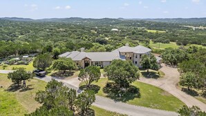A large, single-story house with a spacious driveway and a well-maintained lawn, surrounded by a rural landscape. - Rockin B Summit | Luxury Retreat with Pool & Creek (Pipe Creek)