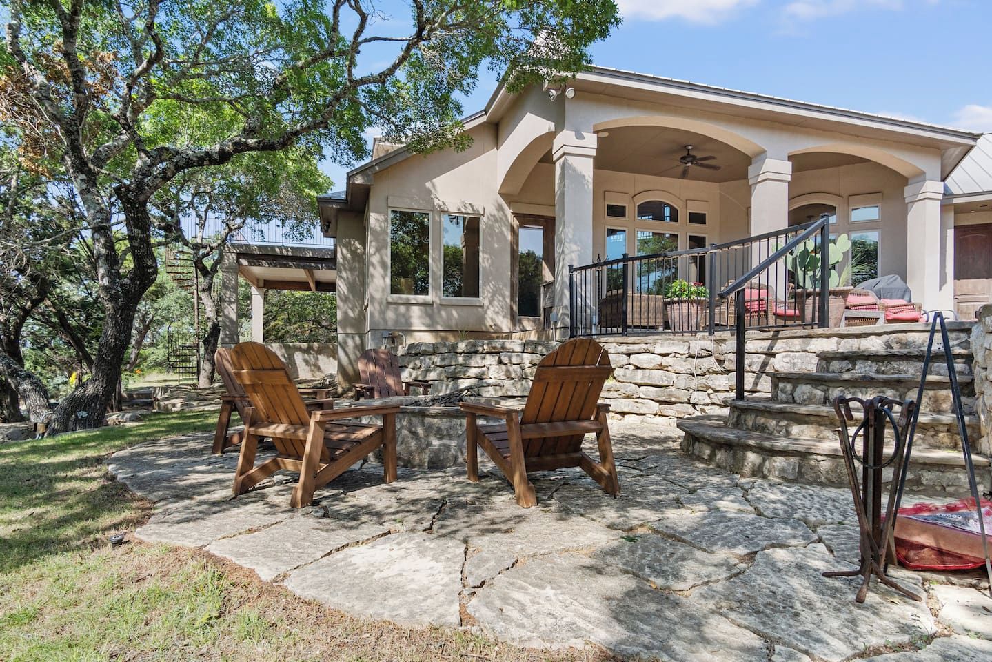 A house with a covered patio area featuring two wooden chairs and a table, stone steps leading to a porch, and a large tree in the background.