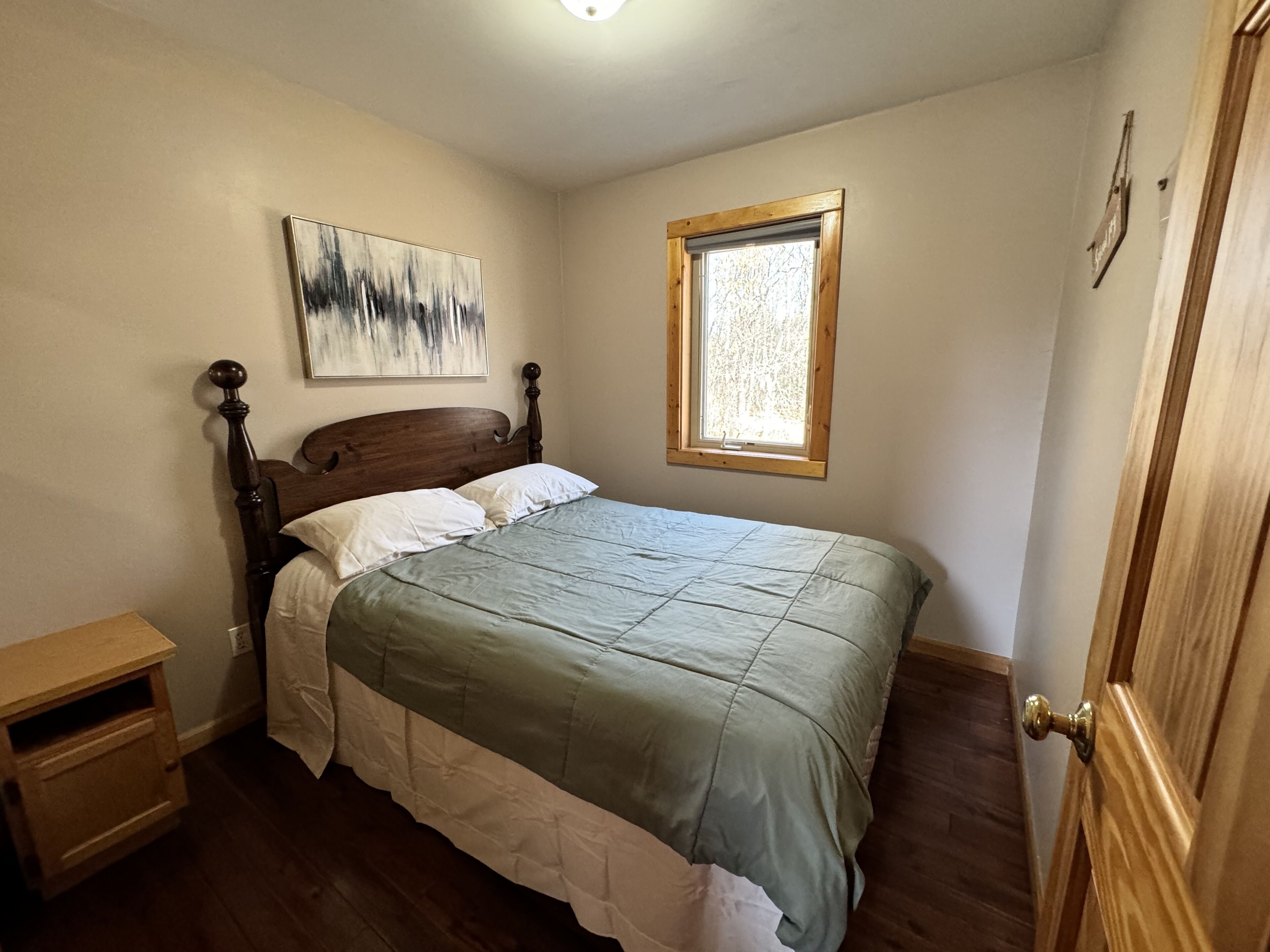 A bedroom with a wooden bed, a nightstand, a window with a view, and a painting on the wall.