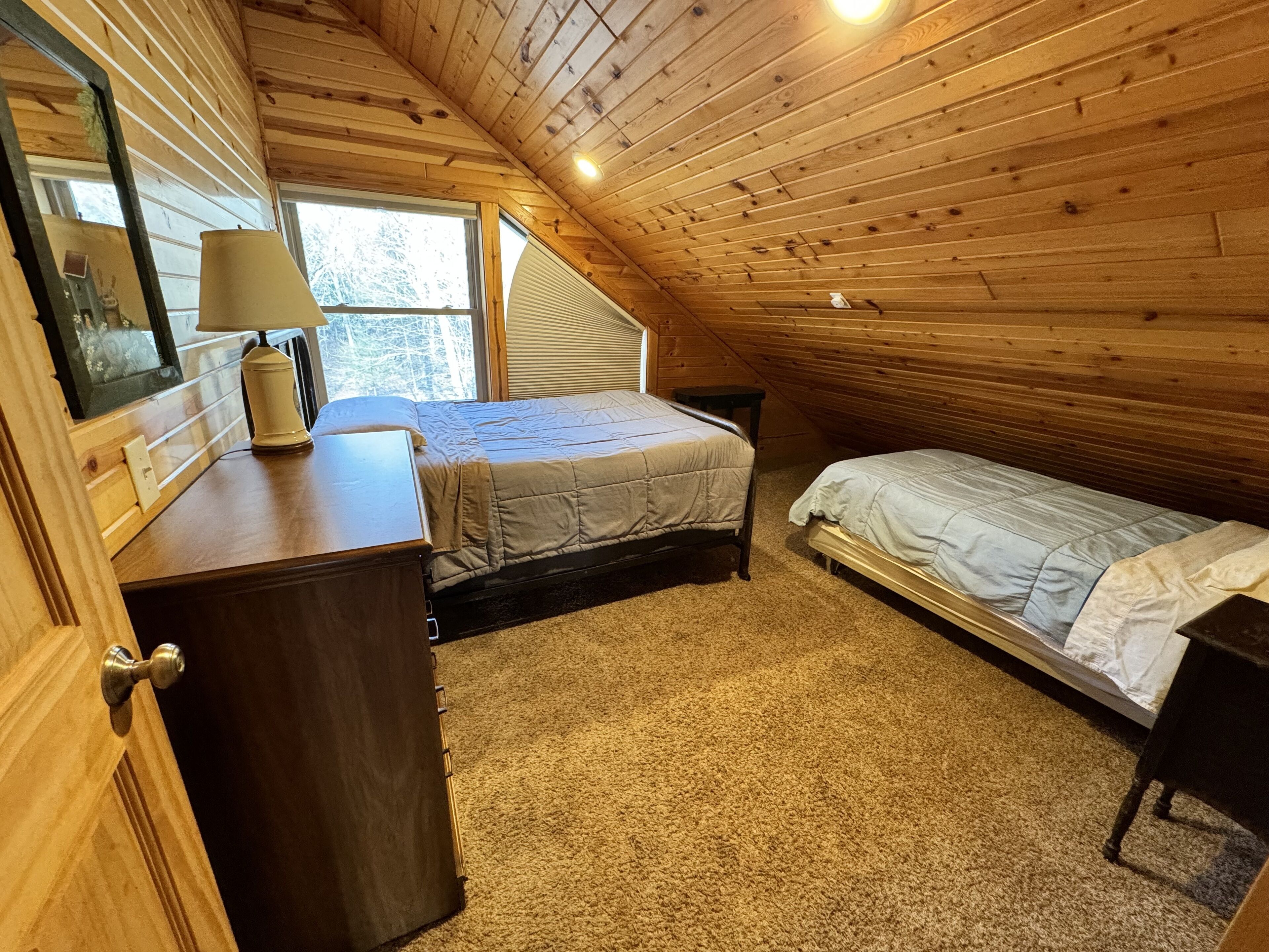 Ein gemütliches Schlafzimmer im Dachgeschoss mit Holzbalkendecke, einem großen Fenster, einem Bett, einem Schreibtisch und einer Kommode.