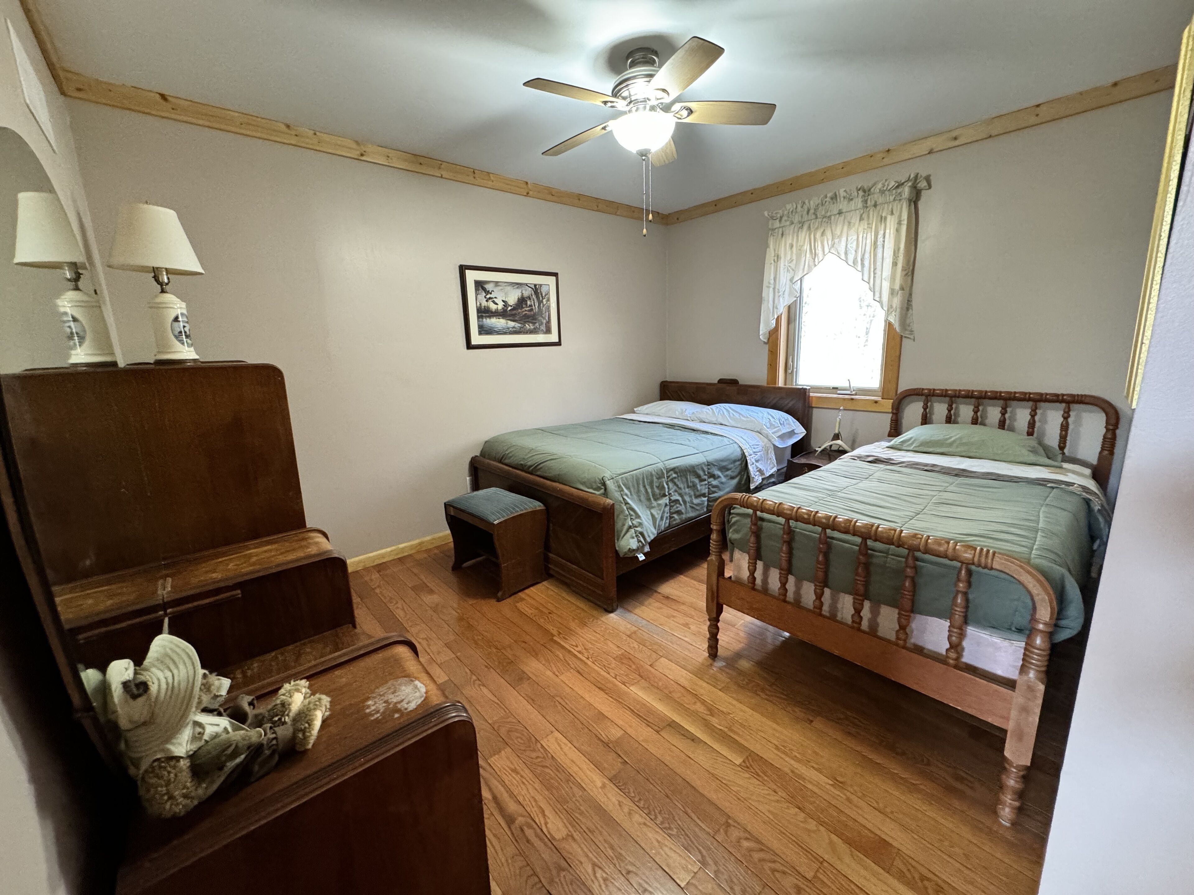 A bedroom with two wooden beds, a ceiling fan, a window with curtains, and a framed picture on the wall.