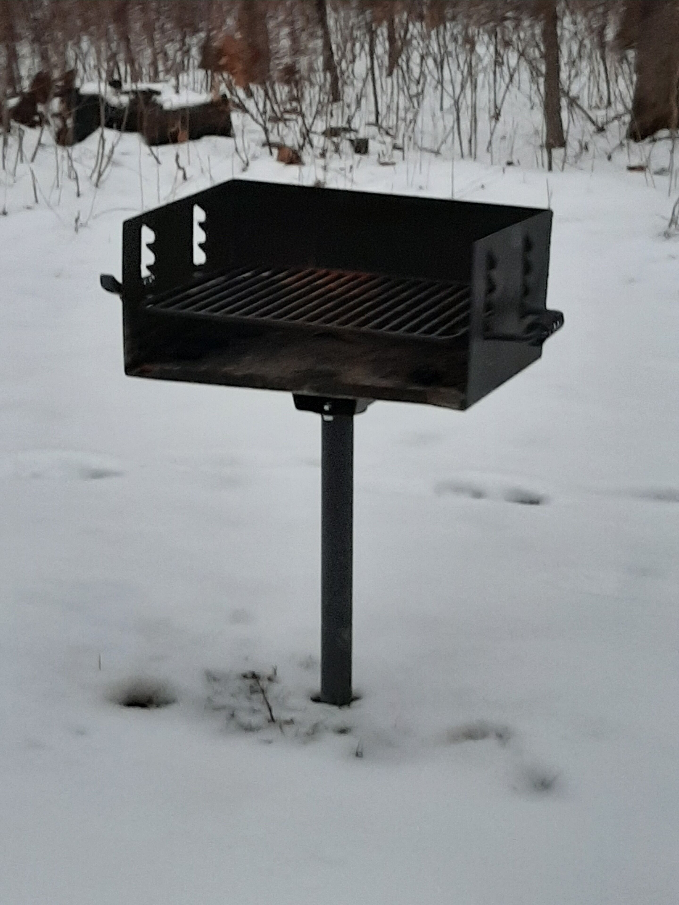 A black barbecue grill with a metal grate, standing on a metal pole in a snowy outdoor setting.