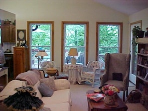 Una sala de estar acogedora con un sofá, sillones, una mesita de centro y una estantería. Tres ventanas ofrecen vistas al exterior.