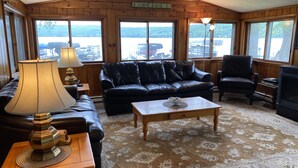 Ein gemütliches Wohnzimmer mit einer Ledersofa, einem Couchtisch und einem Kamin. Große Fenster bieten Blick ins Freie.