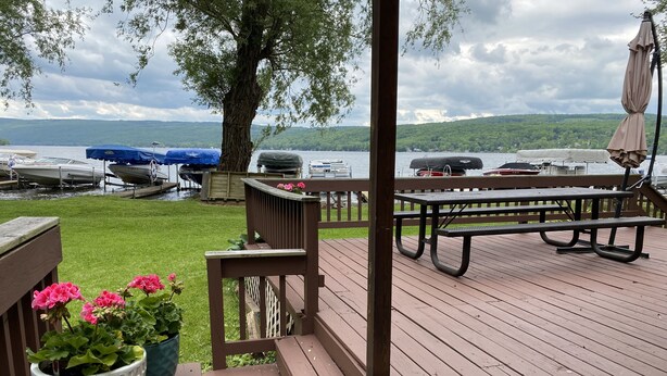 Eine Holzterrasse mit Picknicktischen und Grill, mit Blick auf einen See mit anlegenden Booten.