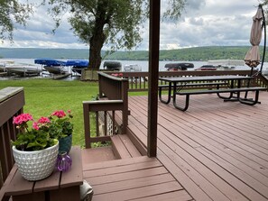 A wooden deck with picnic tables and a grill, overlooking a lake with boats docked. - Eagle Point Cottage on Honeoye Lake (Honeoye)