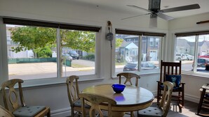 En matplats med ett runt bord och sex trästolar, stora fönster med persienner, en takfläkt och utsikt över en bostadsgata.