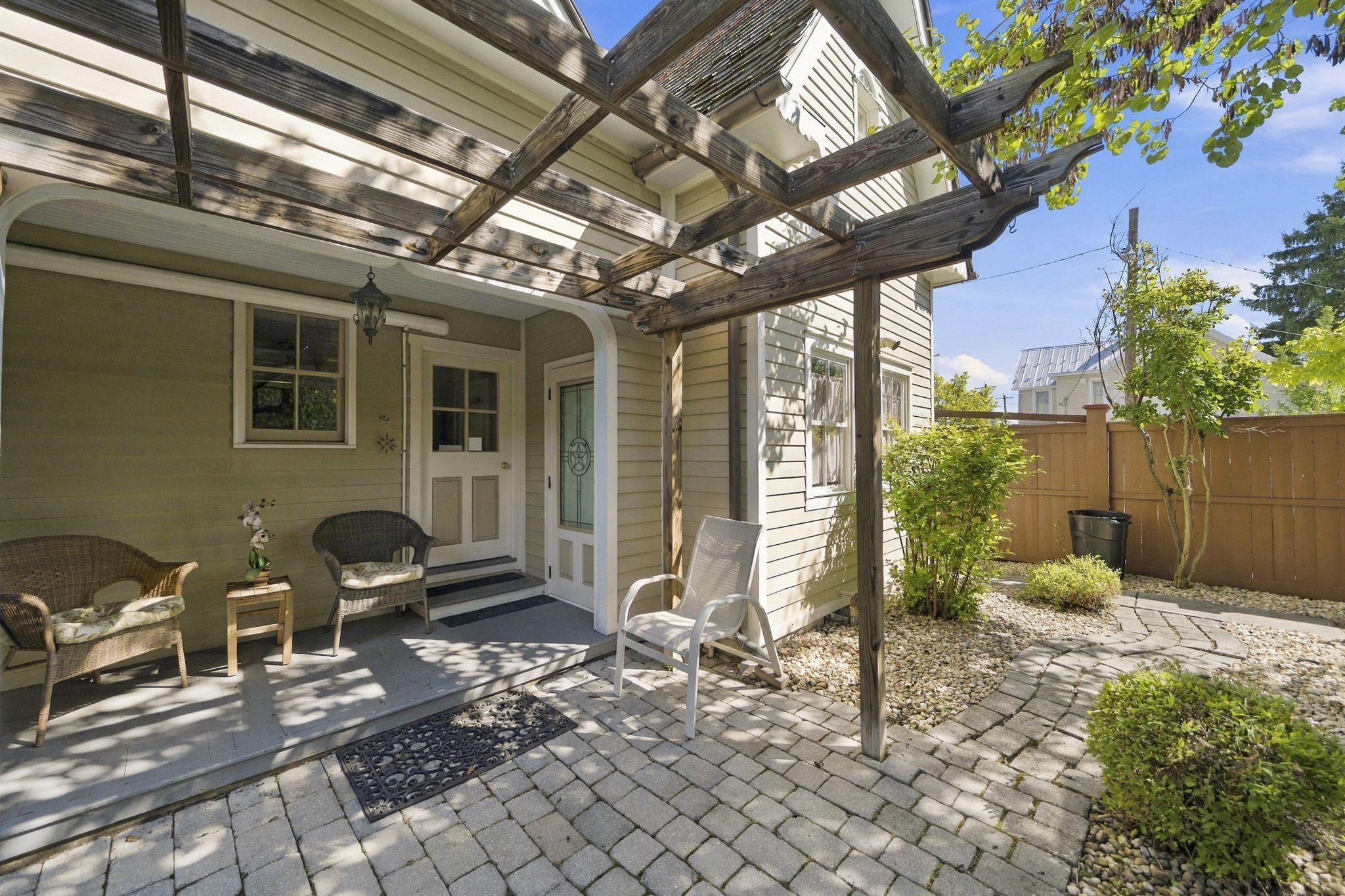 A covered patio with a wooden pergola, a front door, a small porch area with two chairs, and a garden with a wooden fence.