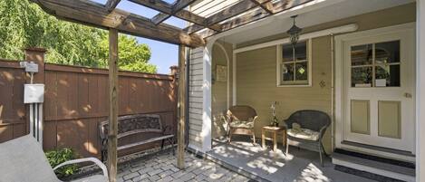 A covered patio with a wooden pergola, outdoor seating, and a door leading to a house.