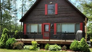 A wooden cabin with a red door and windows, a deck, and surrounded by greenery. - Red Dog Lodge - Beautiful Riverfront Chalet - Sportsperson's Paradise (Grayling)