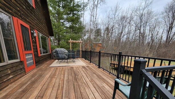 A wooden deck with a table and chairs, a barbecue grill, and a black railing. - Red Dog Lodge - Beautiful Riverfront Chalet - Sportsperson's Paradise (Grayling)