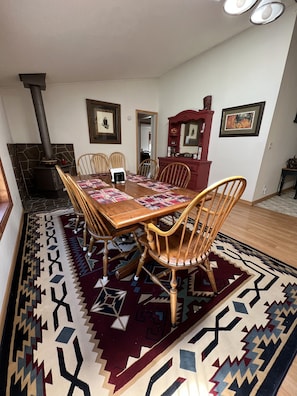 A dining area with a table set for four, surrounded by wooden chairs. A stone fireplace is visible in the background, and the room features a patterned rug and framed artwork on the walls.