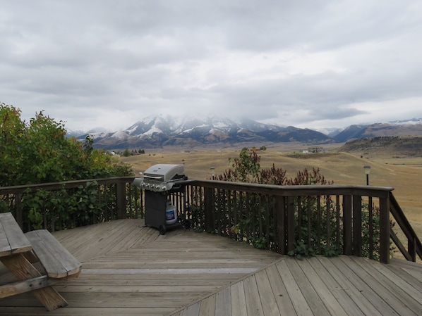 A wooden deck with a barbecue grill and a water bottle, overlooking a mountainous landscape with snow-capped peaks.