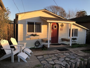 A small white house with a red door, a porch with white Adirondack chairs, and a stone pathway leading to the entrance. - Cozy, Comfortable Beach Home, Steps from the Beach! (Stinson Beach)