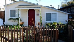 Ein kleines, eingeschossiges Haus mit roter Tür, einer kleinen Veranda und einem Holzzaun.