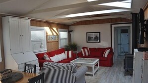 Ein gemütlicher Wohnbereich mit einem roten Sofa, einem gestreiften Sessel und einem weißen Couchtisch. Rechts befindet sich ein weißes Schränkchen und ein Fernseher.