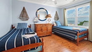 A bedroom with two beds, a wooden dresser, a ceiling fan, a large window with a view of the beach, and decorative wall art. - RARE 6 bedroom, 4 bath OCEANFRONT home, wonderful views & breathtaking sunsets! (Edisto Beach)