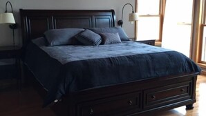 A bedroom with a wooden ceiling, a dark wooden bed frame, a large bed with a dark blue bedspread, two bedside lamps, and a window with a view.