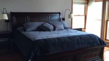Une chambre avec un plafond en bois, un cadre de lit en bois foncé, un grand lit recouvert d’une couverture bleu foncé, deux lampes de chevet et une fenêtre donnant sur un paysage.