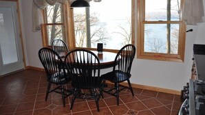 A dining area with a round table and four chairs, large windows with curtains, and a view of trees and water outside.