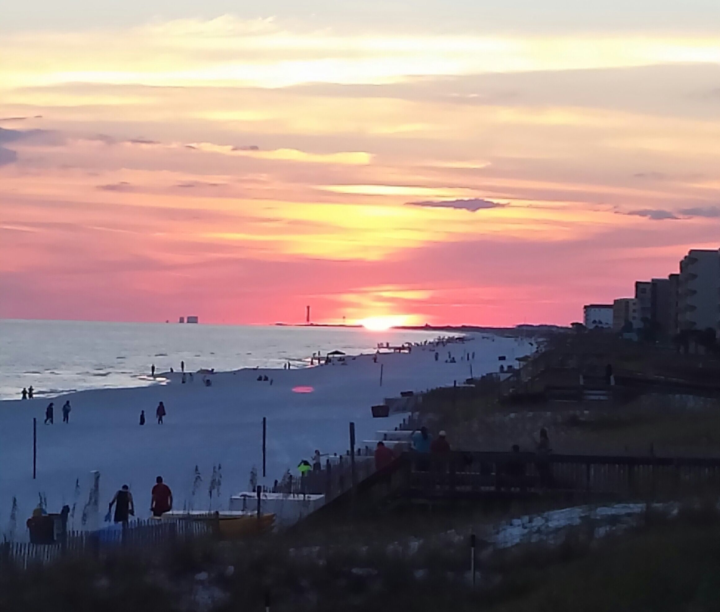 Ein Strand bei Sonnenuntergang, Menschen spazieren am Ufer entlang, im Hintergrund eine Stadtlandschaft.