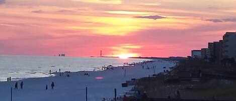 Ein Strand bei Sonnenuntergang, Menschen spazieren am Ufer entlang, im Hintergrund eine Stadtlandschaft.