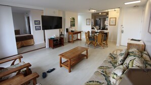 A living room with a wooden coffee table, a sofa with a leaf pattern, a dining area with a wooden table and chairs, and a TV mounted on the wall. - Upgraded 1BR Condo Ocean View - Kitchen, FREE parking, WiFi (Honolulu)