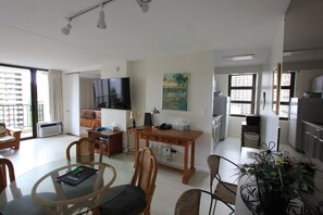 A modern living room with a glass dining table, wicker chairs, a wooden desk, and a flat-screen TV.