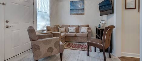 Sala de estar com sofá bege, poltrona estampada, televisão e um quadro pendurado na parede.