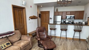 Un salon avec un canapé en cuir marron, un fauteuil inclinable marron et un banc en bois. Il y a également un coin cuisine avec des placards en bois et un réfrigérateur.