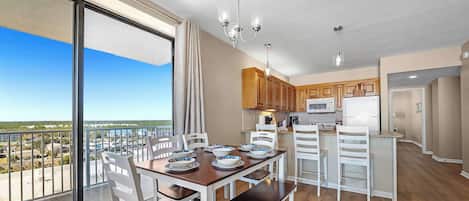 A modern kitchen with wooden cabinets, a dining area with white chairs, and a balcony with a view of the city.