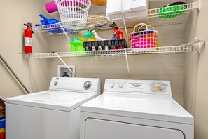 A laundry area with two front-loading washing machines, shelves above holding baskets and cleaning supplies, and a fire extinguisher mounted on the wall.