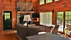 A cozy log cabin interior with a stone fireplace, comfortable seating, and large windows offering views of the outdoors. - Serenity on Cedar-Lakefront Cabin- 10 boats-Swim/Fish/Boat, Starlink & Hulu Live (Tuckasegee)