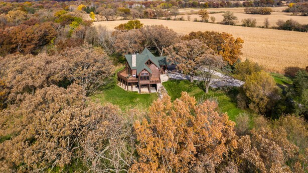 A large, rustic house with a wrap-around porch and a stone foundation, surrounded by autumn-colored trees and open fields.
