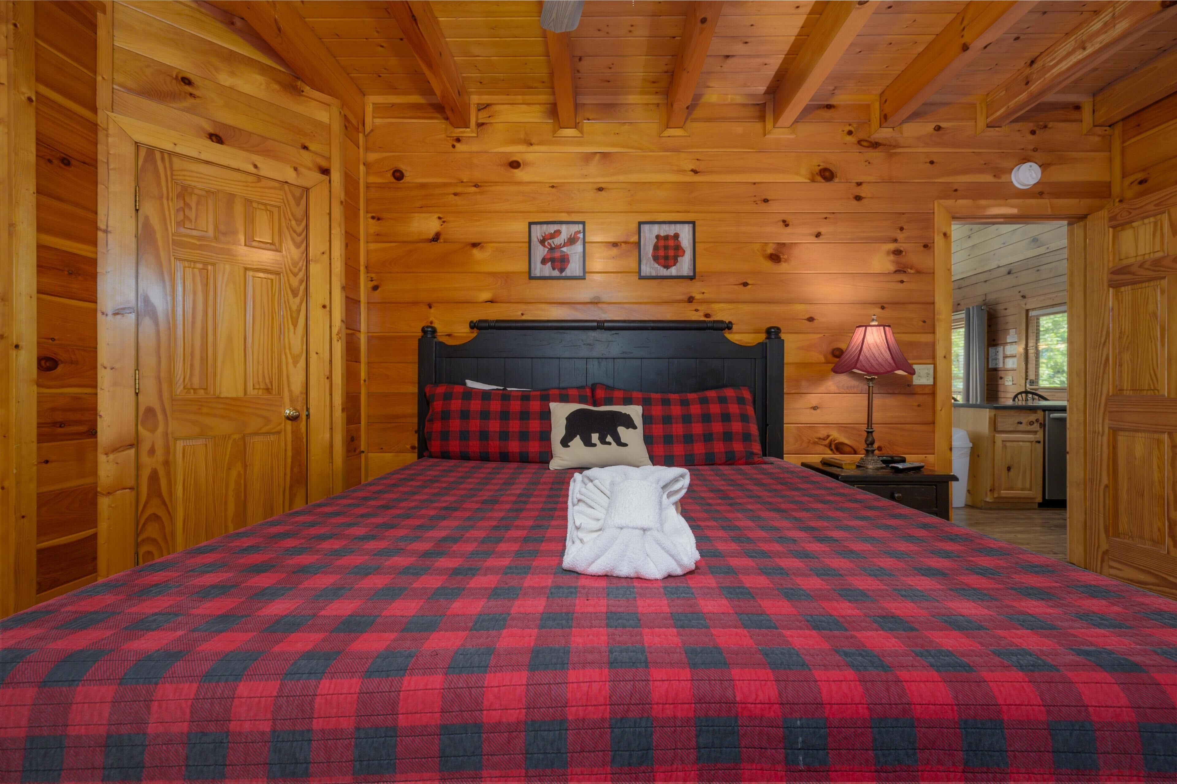 A wooden cabin room with a checkered bedspread, a headboard with a bear pillow, and two framed pictures on the wall.