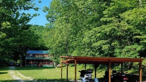Una cabaña rústica con porche y espacio cubierto para vehículos, rodeada de un bosque denso.