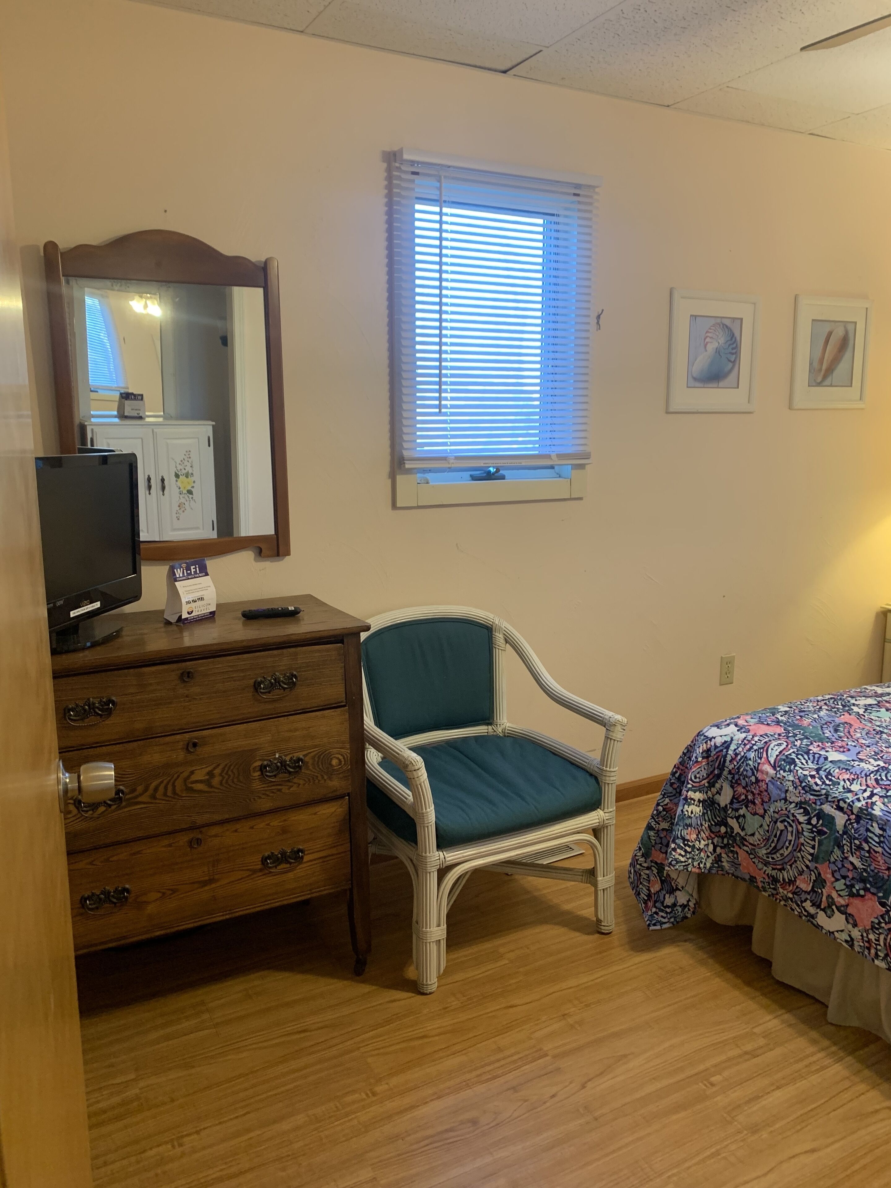 Une chambre avec une commode en bois, un fauteuil en osier, un lit recouvert d’une couvre-lit à motifs floraux, une fenêtre avec des stores et un miroir fixé au mur.