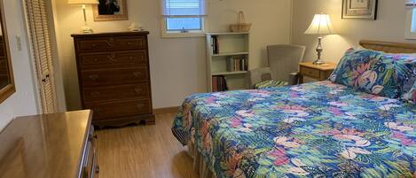 A bedroom with a wooden dresser, a bed with a floral bedspread, a chair, a bookshelf, and a window with blinds.