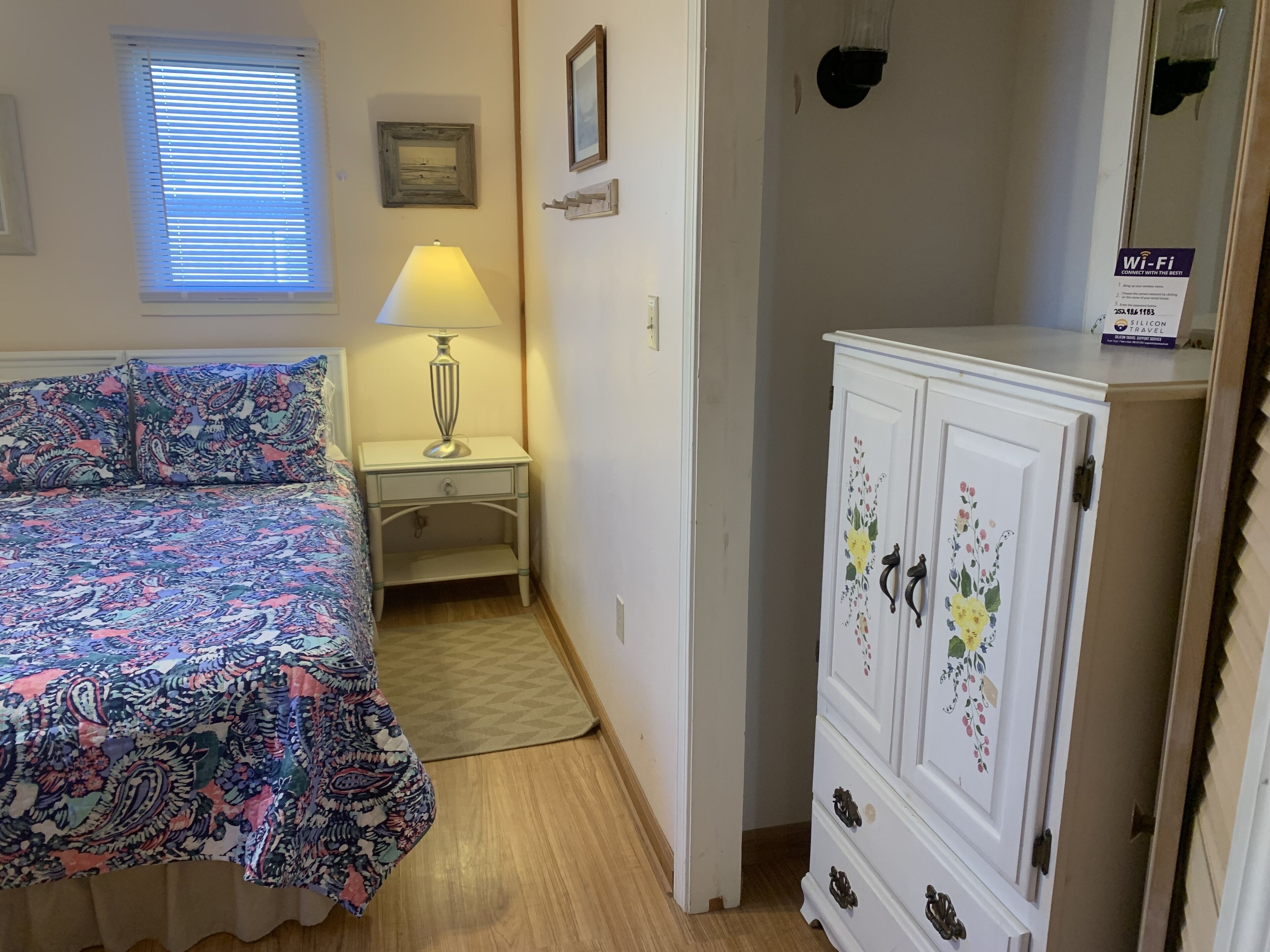 A bedroom with a bed, a nightstand, a lamp, a wardrobe, and a window with blinds.