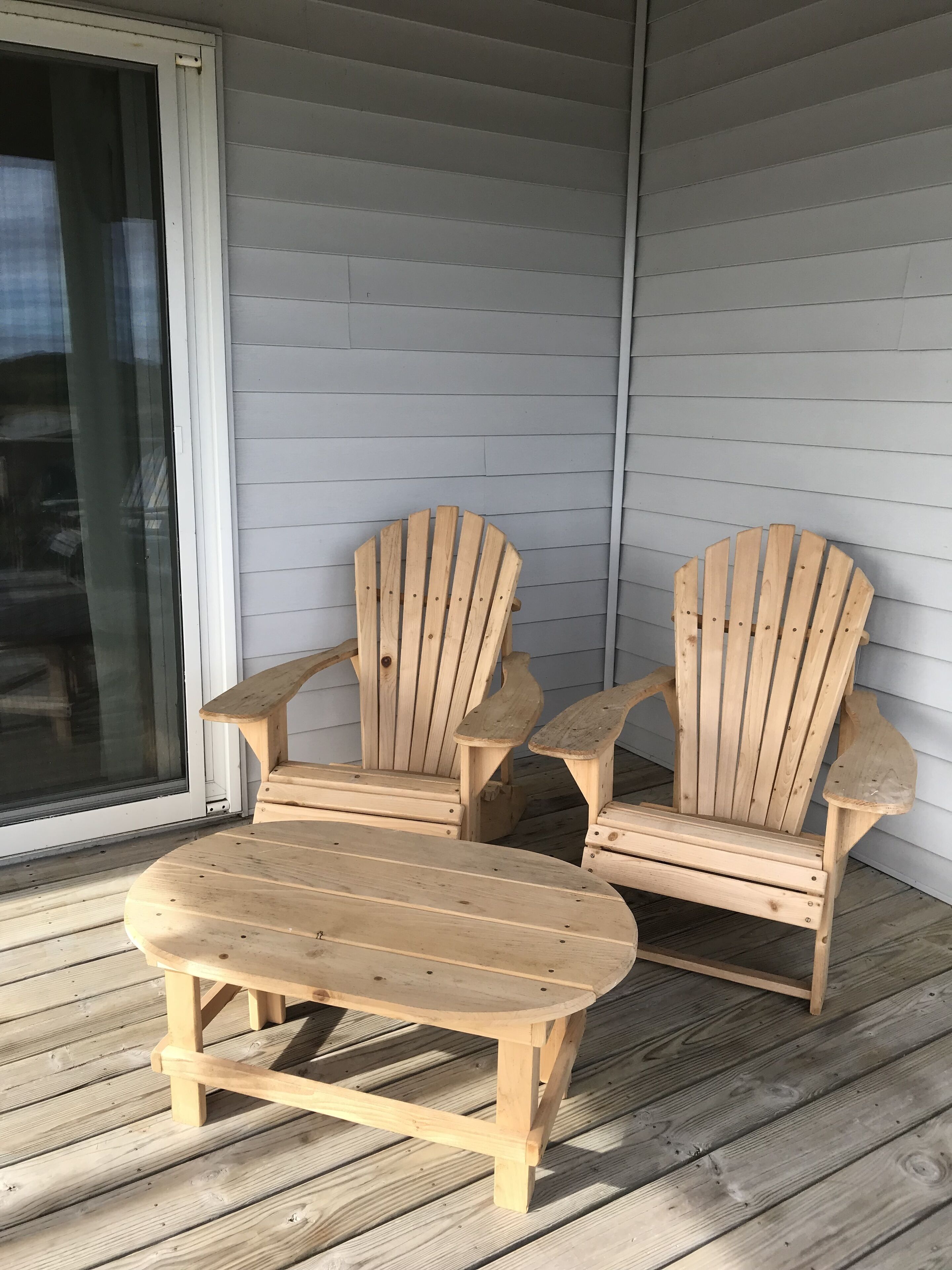 Två trästolar i Adirondack-stil och ett litet bord står på ett trädäck, med en skjutdörr i glas som syns i bakgrunden.