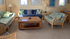 A living room with a blue sofa, a wooden coffee table, and wicker furniture. There are two lamps, a framed fish poster, and a painting on the wall. - Frisco Beach House! Some ocean views! Easy walk to the beach!  (Frisco)